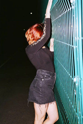 Young redhead with short black skirt standing facing an industrial fence